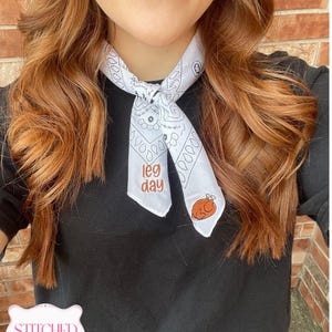 May include: A white bandana featuring a black paisley pattern, the embroidered text "leg day" in orange, and an embroidered graphic of a roasted turkey. The bandana is tied around the neck over a plain black shirt.