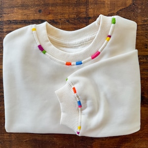 May include: A white sweatshirt with a colorful trim around the neckline and the cuffs. The trim is made of different colored threads, including red, orange, yellow, green, blue, and purple.