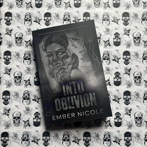 May include: A paperback book titled Into Oblivion by Ember Nicole, featuring a grayscale illustration of a person with snakes and a chain. The cover text reads A Modernized Retelling of the Tale of Medusa. The book rests on a white background with a repeating pattern of black skulls and roses.