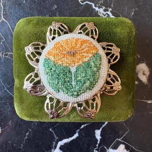 Puede incluir: Broche redondo bordado con un dise&ntilde;o floral en tonos naranja y verde. La flor bordada est&aacute; engastada en un marco met&aacute;lico calado de color plateado. El broche descansa sobre una superficie cuadrada de terciopelo verde.