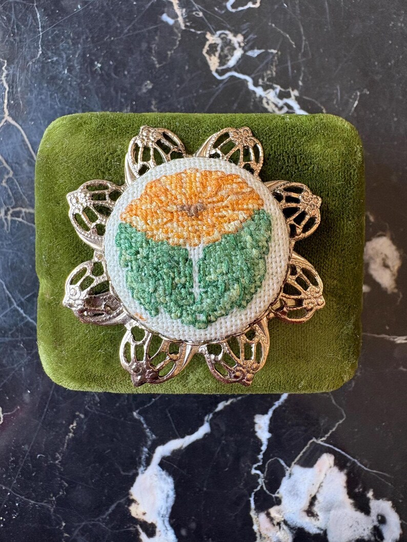 Puede incluir: Broche redondo bordado con un dise&ntilde;o floral en tonos naranja y verde. La flor bordada est&aacute; engastada en un marco met&aacute;lico calado de color plateado. El broche descansa sobre una superficie cuadrada de terciopelo verde.