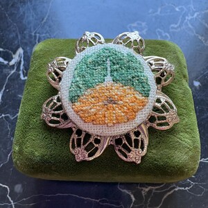 Puede incluir: Broche de metal plateado con dise&ntilde;o floral. El centro presenta una imagen bordada a punto de cruz de un edificio con una combinaci&oacute;n de colores verde y naranja. El broche est&aacute; sobre una superficie de terciopelo verde.