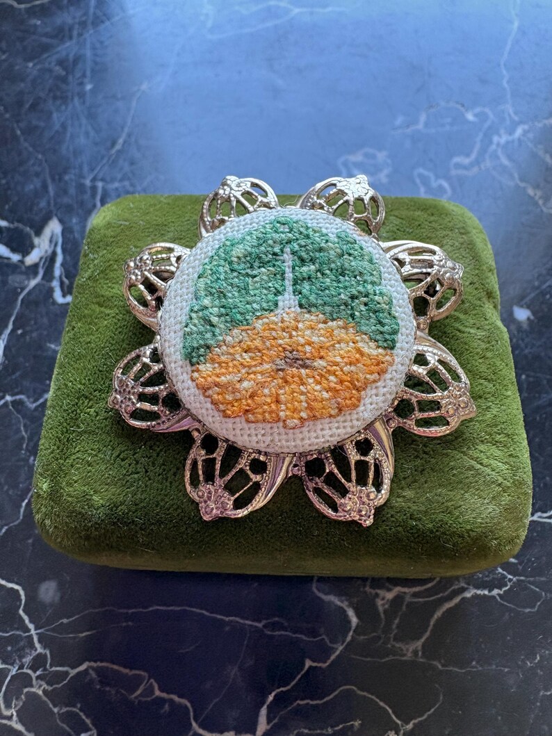 Puede incluir: Broche de metal plateado con dise&ntilde;o floral. El centro presenta una imagen bordada a punto de cruz de un edificio con una combinaci&oacute;n de colores verde y naranja. El broche est&aacute; sobre una superficie de terciopelo verde.