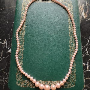 May include: A delicate pink pearl necklace with a decorative silver clasp. The necklace is displayed on a vintage green jewelry box with gold filigree detailing. The pearls graduate in size, creating an elegant look.