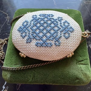Puede incluir: Colgante ovalado de color crema con un dise&ntilde;o de punto de cruz azul. El colgante est&aacute; sujeto a una cadena dorada y descansa sobre una superficie de terciopelo verde. El colgante tiene detalles met&aacute;licos dorados.