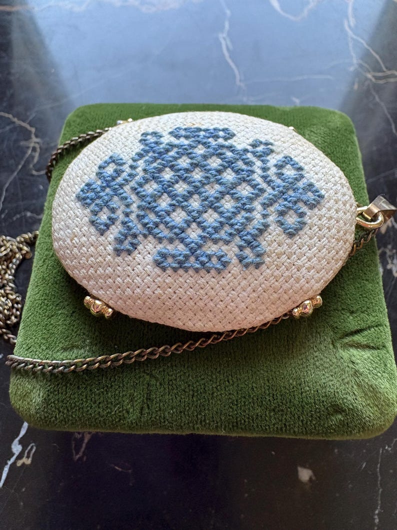 Puede incluir: Colgante ovalado de color crema con un dise&ntilde;o de punto de cruz azul. El colgante est&aacute; sujeto a una cadena dorada y descansa sobre una superficie de terciopelo verde. El colgante tiene detalles met&aacute;licos dorados.