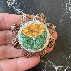 Puede incluir: Broche floral de estilo vintage. El broche presenta una flor bordada detallada en tonos amarillos y verdes, engastada en un marco de filigrana dorada. El dise&ntilde;o es circular, con la flor en el centro y el marco en forma de flor estilizada.