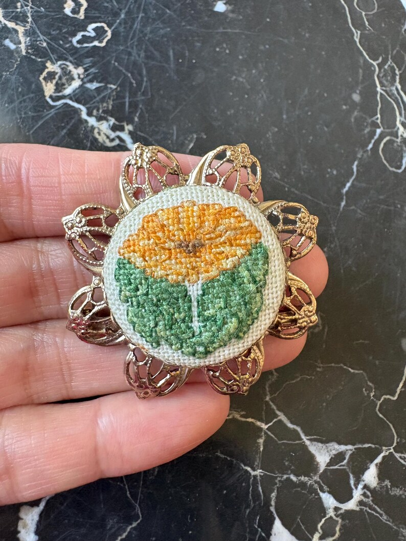 Puede incluir: Broche floral de estilo vintage. El broche presenta una flor bordada detallada en tonos amarillos y verdes, engastada en un marco de filigrana dorada. El dise&ntilde;o es circular, con la flor en el centro y el marco en forma de flor estilizada.