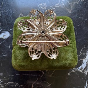 Puede incluir: Broche de flores caladas en tono dorado con cierre de alfiler. El broche descansa sobre un peque&ntilde;o coj&iacute;n de terciopelo verde. El dise&ntilde;o floral presenta detalles intrincados y un acento negro central.