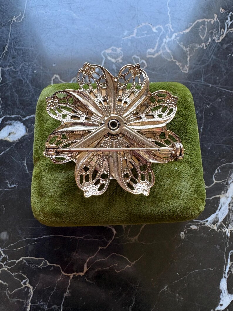 Puede incluir: Broche de flores caladas en tono dorado con cierre de alfiler. El broche descansa sobre un peque&ntilde;o coj&iacute;n de terciopelo verde. El dise&ntilde;o floral presenta detalles intrincados y un acento negro central.