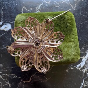 Puede incluir: Broche de flores plateada vintage con un intrincado dise&ntilde;o de p&eacute;talos calados. La broche est&aacute; colocada sobre una superficie de terciopelo verde, mostrando su detallada artesan&iacute;a. El alfiler es visible, listo para usar.
