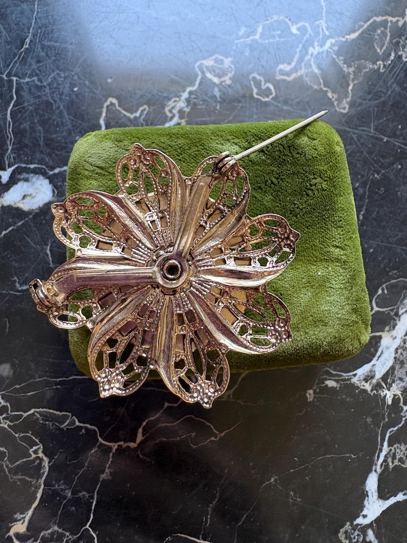 Puede incluir: Broche de flores plateada vintage con un intrincado dise&ntilde;o de p&eacute;talos calados. La broche est&aacute; colocada sobre una superficie de terciopelo verde, mostrando su detallada artesan&iacute;a. El alfiler es visible, listo para usar.