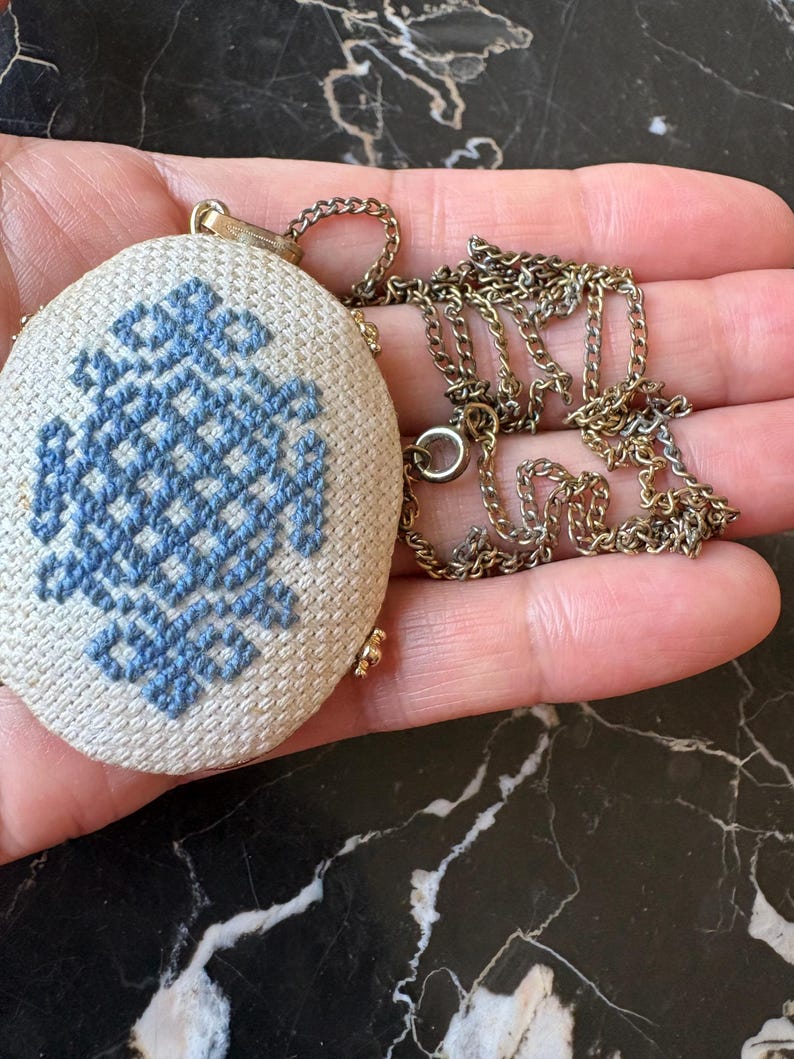 Puede incluir: Collar con colgante ovalado con base de tela color crema y dise&ntilde;o de punto de cruz azul. El colgante est&aacute; sujeto a una cadena dorada. El collar se sostiene en una mano.