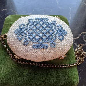 Puede incluir: Un bolso ovalado vintage con una parte superior de punto de cruz crema con un dise&ntilde;o floral azul. Tiene una correa de cadena dorada y est&aacute; colocado sobre una superficie de terciopelo verde. Los bordes del bolso est&aacute;n adornados con metal dorado.