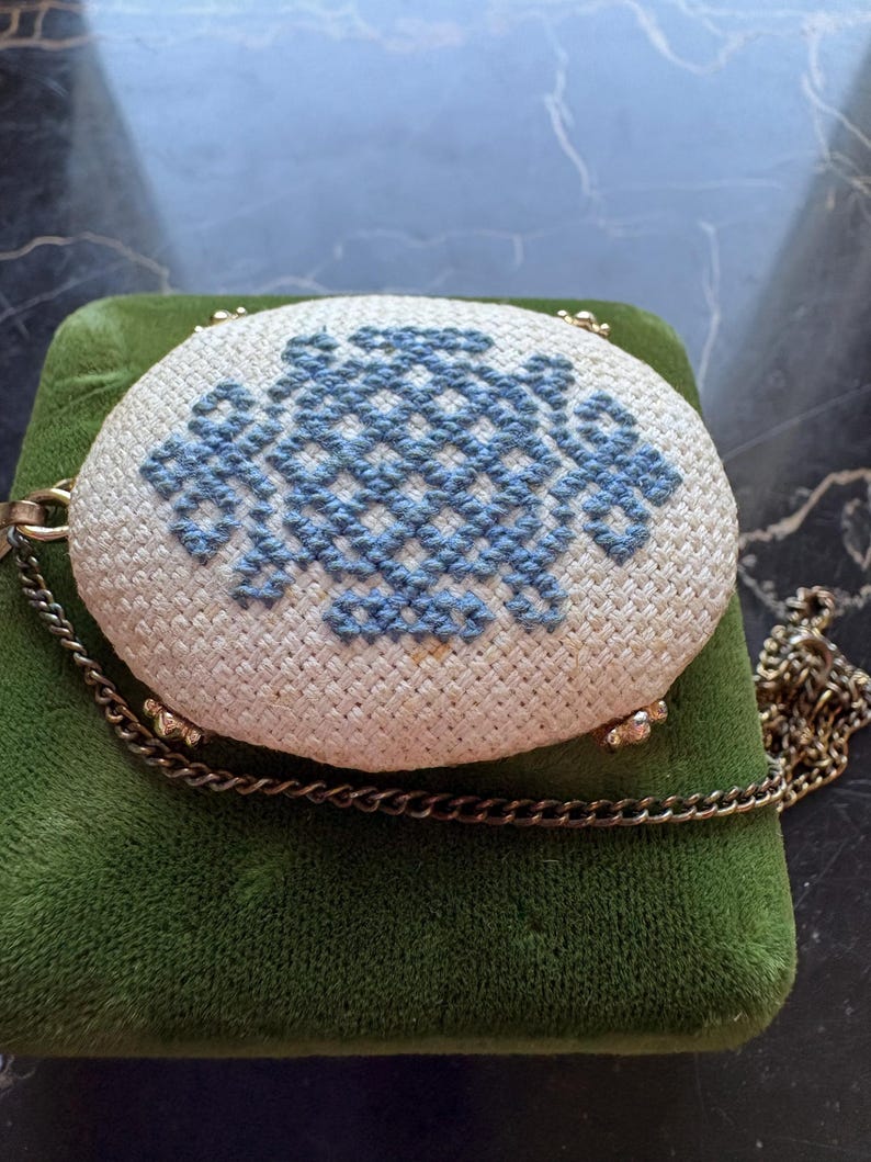 Puede incluir: Un bolso ovalado vintage con una parte superior de punto de cruz crema con un dise&ntilde;o floral azul. Tiene una correa de cadena dorada y est&aacute; colocado sobre una superficie de terciopelo verde. Los bordes del bolso est&aacute;n adornados con metal dorado.