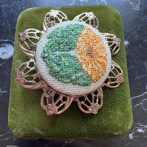 Puede incluir: Broche de estilo vintage con dise&ntilde;o floral. El broche presenta un c&iacute;rculo central bordado con secciones verdes y naranjas, engastado en un marco de metal calado plateado. El broche se exhibe sobre una superficie de terciopelo verde.