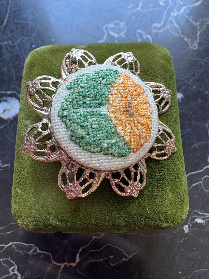 Puede incluir: Broche de estilo vintage con dise&ntilde;o floral. El broche presenta un c&iacute;rculo central bordado con secciones verdes y naranjas, engastado en un marco de metal calado plateado. El broche se exhibe sobre una superficie de terciopelo verde.