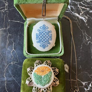 Puede incluir: Un collar con colgante vintage con un dise&ntilde;o bordado en azul y blanco, exhibido en una caja de joyer&iacute;a de terciopelo verde. Tambi&eacute;n se ve un broche bordado a juego con un dise&ntilde;o floral.
