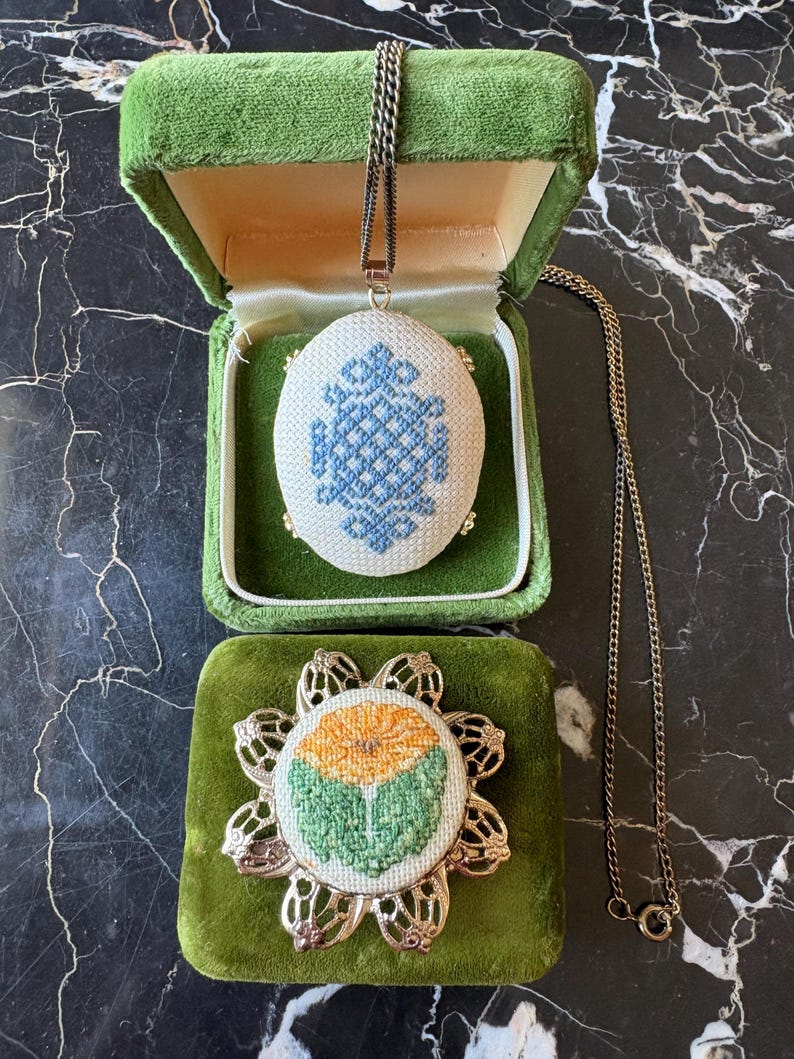 Puede incluir: Un collar con colgante vintage con un dise&ntilde;o bordado en azul y blanco, exhibido en una caja de joyer&iacute;a de terciopelo verde. Tambi&eacute;n se ve un broche bordado a juego con un dise&ntilde;o floral.