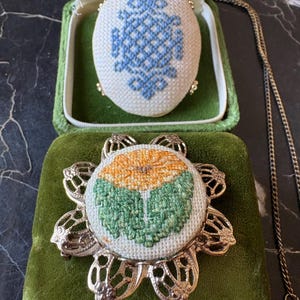 Puede incluir: Un conjunto de joyas vintage con un colgante y un broche. El colgante es de forma ovalada con un dise&ntilde;o de punto de cruz azul, presentado en una caja de terciopelo verde. El broche tiene un dise&ntilde;o floral con punto de cruz naranja y verde, engastado en un marco de metal dorado.
