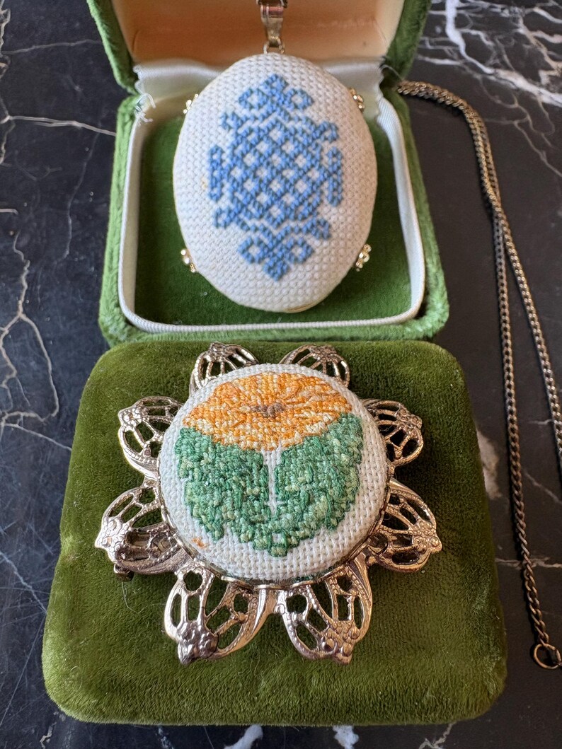 Puede incluir: Un conjunto de joyas vintage con un colgante y un broche. El colgante es de forma ovalada con un dise&ntilde;o de punto de cruz azul, presentado en una caja de terciopelo verde. El broche tiene un dise&ntilde;o floral con punto de cruz naranja y verde, engastado en un marco de metal dorado.