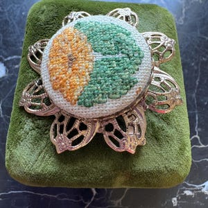 Puede incluir: Un coj&iacute;n redondo bordado con un dise&ntilde;o floral en naranja y verde, colocado sobre un coj&iacute;n de terciopelo verde. El coj&iacute;n est&aacute; rodeado por un marco decorativo de metal plateado.