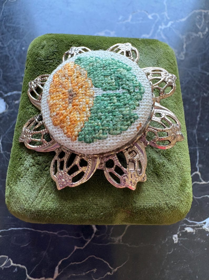 Puede incluir: Un coj&iacute;n redondo bordado con un dise&ntilde;o floral en naranja y verde, colocado sobre un coj&iacute;n de terciopelo verde. El coj&iacute;n est&aacute; rodeado por un marco decorativo de metal plateado.