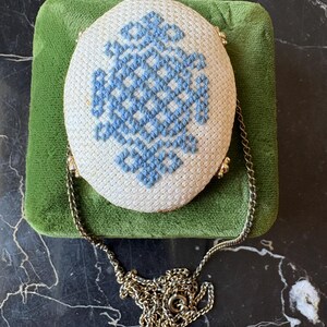 Puede incluir: Bolso ovalado blanco con dise&ntilde;o de punto de cruz azul. Cuenta con una cadena y cierre dorados. El bolso descansa sobre una superficie de terciopelo verde, con un aspecto vintage.