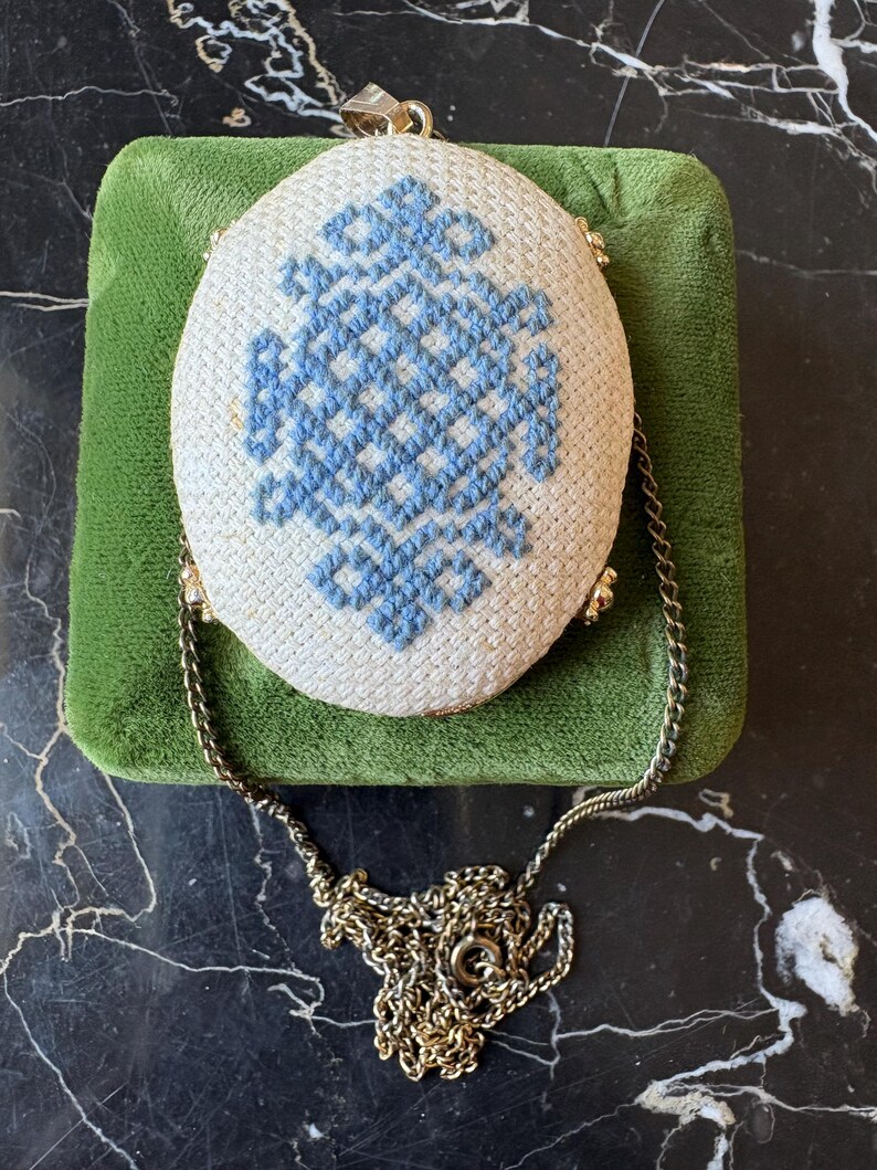 Puede incluir: Bolso ovalado blanco con dise&ntilde;o de punto de cruz azul. Cuenta con una cadena y cierre dorados. El bolso descansa sobre una superficie de terciopelo verde, con un aspecto vintage.