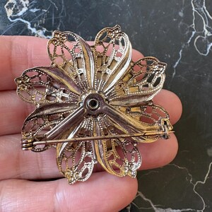 Puede incluir: Broche de flor de metal dorado vintage con intrincados detalles de filigrana. El broche tiene un cierre de alfiler en la parte posterior. El dise&ntilde;o floral presenta m&uacute;ltiples p&eacute;talos con patrones calados, creando una apariencia delicada y ornamentada.