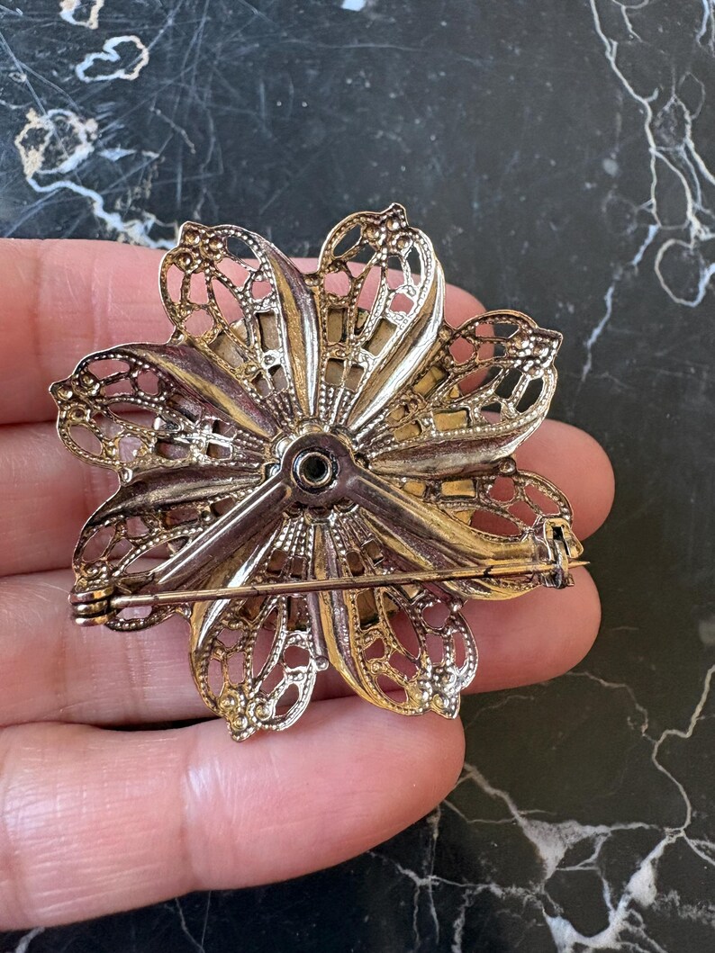 Puede incluir: Broche de flor de metal dorado vintage con intrincados detalles de filigrana. El broche tiene un cierre de alfiler en la parte posterior. El dise&ntilde;o floral presenta m&uacute;ltiples p&eacute;talos con patrones calados, creando una apariencia delicada y ornamentada.
