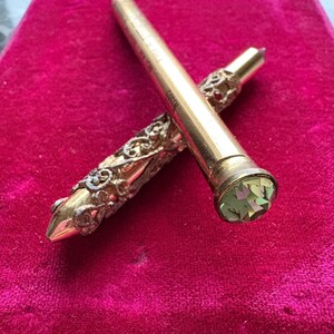 May include: A gold-toned mechanical pencil featuring an intricate filigree metal casing and a faceted light green gemstone set into the top end. The barrel is smooth with visible text and the tip is pointed, showcasing a detailed metal design.