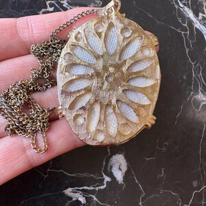 Puede incluir: Collar con colgante ovalado dorado con dise&ntilde;o floral. El colgante presenta un centro texturizado blanquecino con un marco y cadena dorados. El collar se muestra sobre una superficie oscura y veteada.