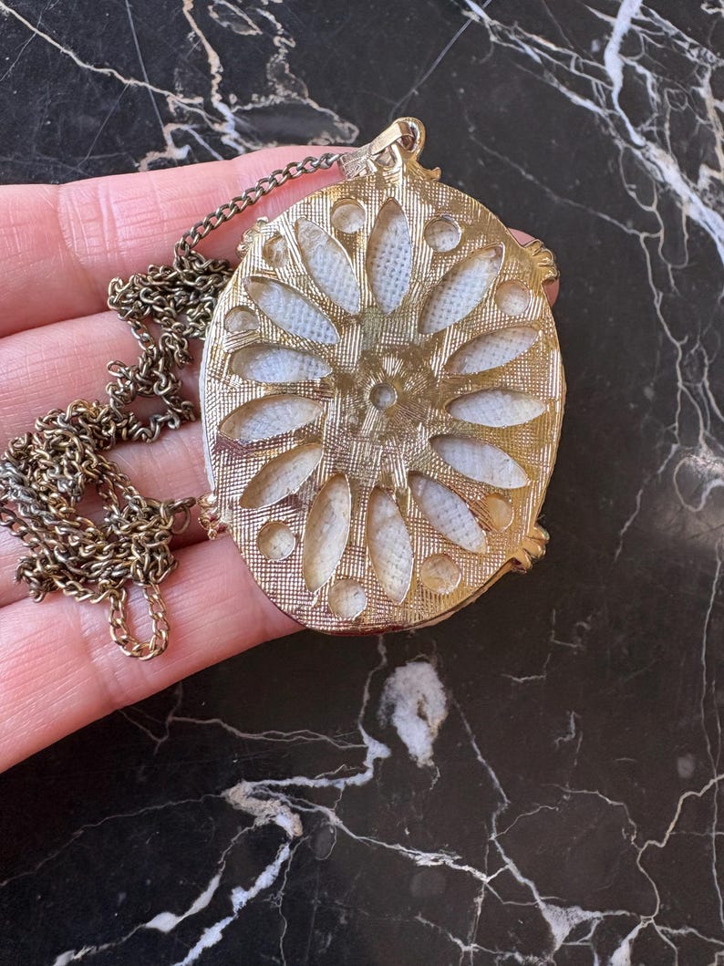 Puede incluir: Collar con colgante ovalado dorado con dise&ntilde;o floral. El colgante presenta un centro texturizado blanquecino con un marco y cadena dorados. El collar se muestra sobre una superficie oscura y veteada.