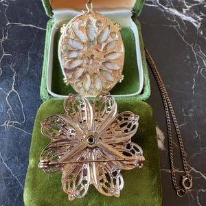 Puede incluir: Un conjunto de joyas vintage en tono dorado con un collar con colgante y un broche en forma de flor. El colgante es ovalado con un dise&ntilde;o floral, y el broche tiene un dise&ntilde;o de p&eacute;talos calados. Ambas piezas descansan sobre una superficie de terciopelo verde.