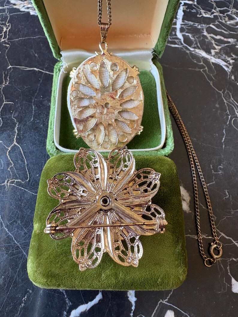 Puede incluir: Un conjunto de joyas vintage en tono dorado con un collar con colgante y un broche en forma de flor. El colgante es ovalado con un dise&ntilde;o floral, y el broche tiene un dise&ntilde;o de p&eacute;talos calados. Ambas piezas descansan sobre una superficie de terciopelo verde.