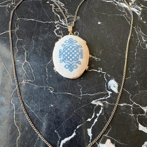 Puede incluir: Un collar dorado con un colgante ovalado con un dise&ntilde;o bordado en azul y blanco. La fina cadena y el colgante crean un estilo vintage. Se muestra sobre una superficie de m&aacute;rmol negro.