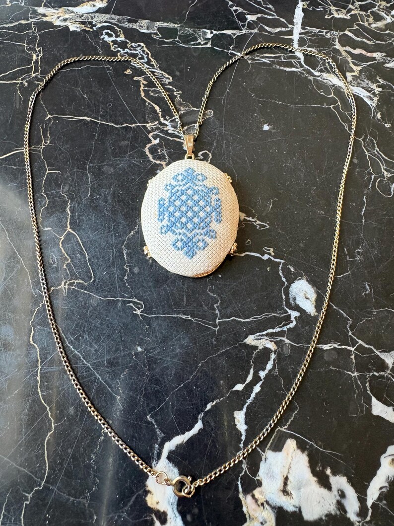 Puede incluir: Un collar dorado con un colgante ovalado con un dise&ntilde;o bordado en azul y blanco. La fina cadena y el colgante crean un estilo vintage. Se muestra sobre una superficie de m&aacute;rmol negro.