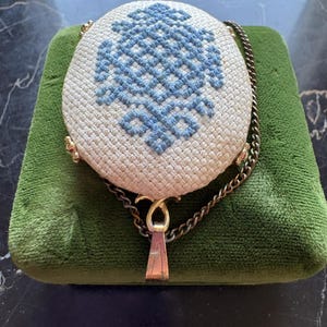 Puede incluir: Un bolso vintage redondo de color crema con un dise&ntilde;o bordado en azul. El bolso tiene un cierre dorado y una correa de cadena. El bolso est&aacute; sobre una superficie de terciopelo verde.