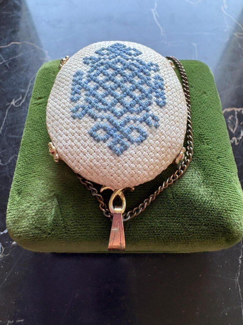 Puede incluir: Un bolso vintage redondo de color crema con un dise&ntilde;o bordado en azul. El bolso tiene un cierre dorado y una correa de cadena. El bolso est&aacute; sobre una superficie de terciopelo verde.