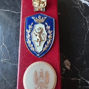 May include: Three metal pins displayed on a red rectangular base. The top pin is a gold shield with a lion and the text DIEU ET MON DROIT. The middle pin is a blue shield with a gold lion, crown, and laurel. The bottom pin is a round gold eagle emblem.