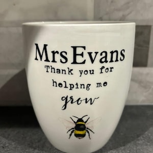 May include: White ceramic mug with the text "Mrs Evans Thank you for helping me grow" in black font. A yellow and black bee illustration is below the text. The mug is on a grey surface.