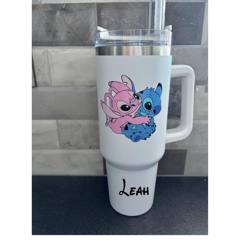 Puede incluir: Vaso t&eacute;rmico blanco con asa, con una imagen de dibujos animados de Stitch y Angel. El vaso tiene una tapa transparente y una pajita. El nombre "Leah" est&aacute; impreso en negro en la parte inferior.