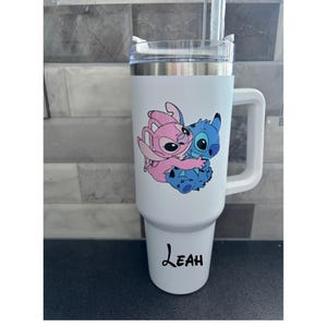 Puede incluir: Vaso t&eacute;rmico blanco con asa, con una imagen de dibujos animados de Stitch y Angel abraz&aacute;ndose. El vaso tiene una tapa transparente y una pajita. El nombre "Leah" est&aacute; impreso en escritura negra en la parte inferior.