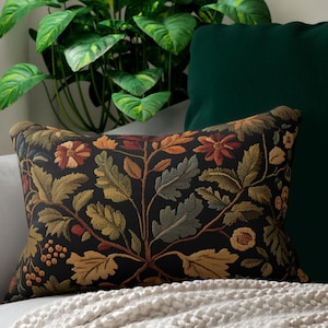 Op de afbeelding: Een decoratief rechthoekig kussen met een zwarte achtergrond en een bloemenpatroon in groen, oranje en bruin. Het ligt op een grijze bank met een crèmekleurige deken. Een groen kussen en een kamerplant zijn op de achtergrond. Afmetingen: ongeveer 40 x 60 cm.