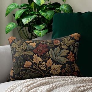 May include: Rectangular cushion featuring a dark background with a floral design in green, orange, and red hues. The cushion is placed on a light grey sofa, with a cream-coloured blanket.