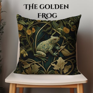 May include: Square cushion with a detailed illustration of a green frog amidst stylized foliage in shades of green, gold, and dark blue. The text "THE GOLDEN FROG" is arched across the top. The cushion is placed on a white chair.