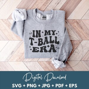 May include: A gray sweatshirt with the text "IN MY T-BALL ERA" in black lettering with a sparkly design. The text is in a retro font style.