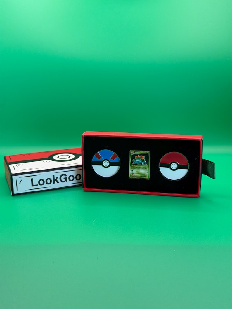 May include: A red and black box set featuring two Pok&eacute; Ball pins and a Pok&eacute;mon trading card. The box has the text "LookGoo" on the side. The background is a solid green.