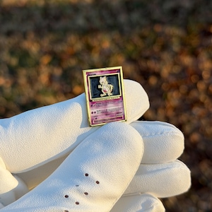 May include: A gold-framed pin featuring a Pokemon card design with a purple and pink Mewtwo illustration. The pin is held by a person wearing a white glove. The background is blurred with natural tones.